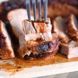 Fork piercing stack of two pieces of cooked pork tenderloin on wood cutting board.
