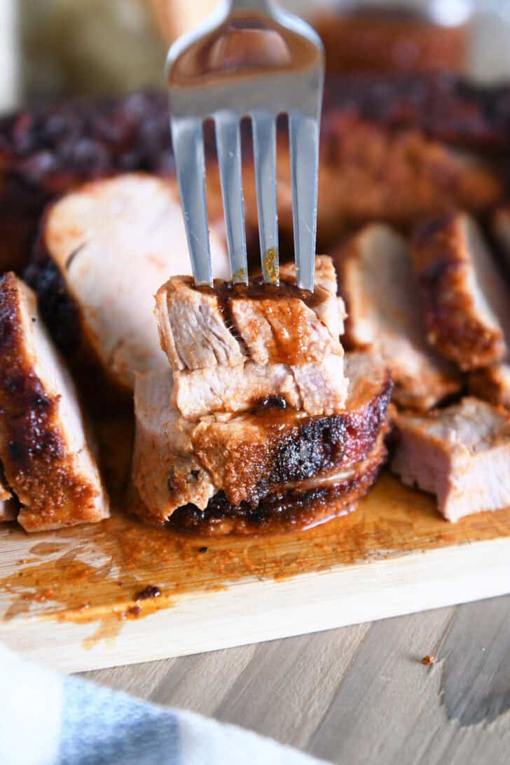 Fork piercing two stacked pieces of cooked pork tenderloin on wood cutting board.