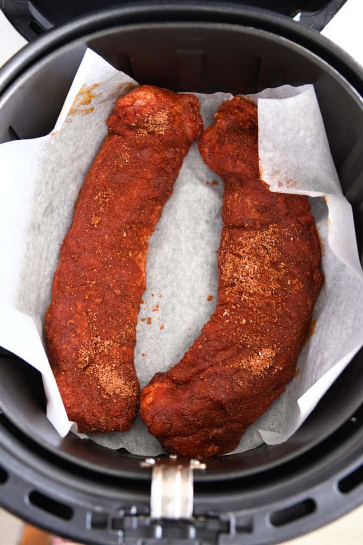 Two spice-crusted uncooked pork tenderloins on parchment paper in air fryer.