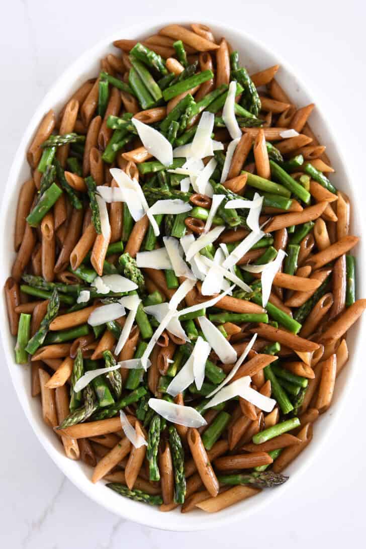 Top down view of penne pasta in white serving dish with roasted asapragus, balsamic butter and parmesan cheese.