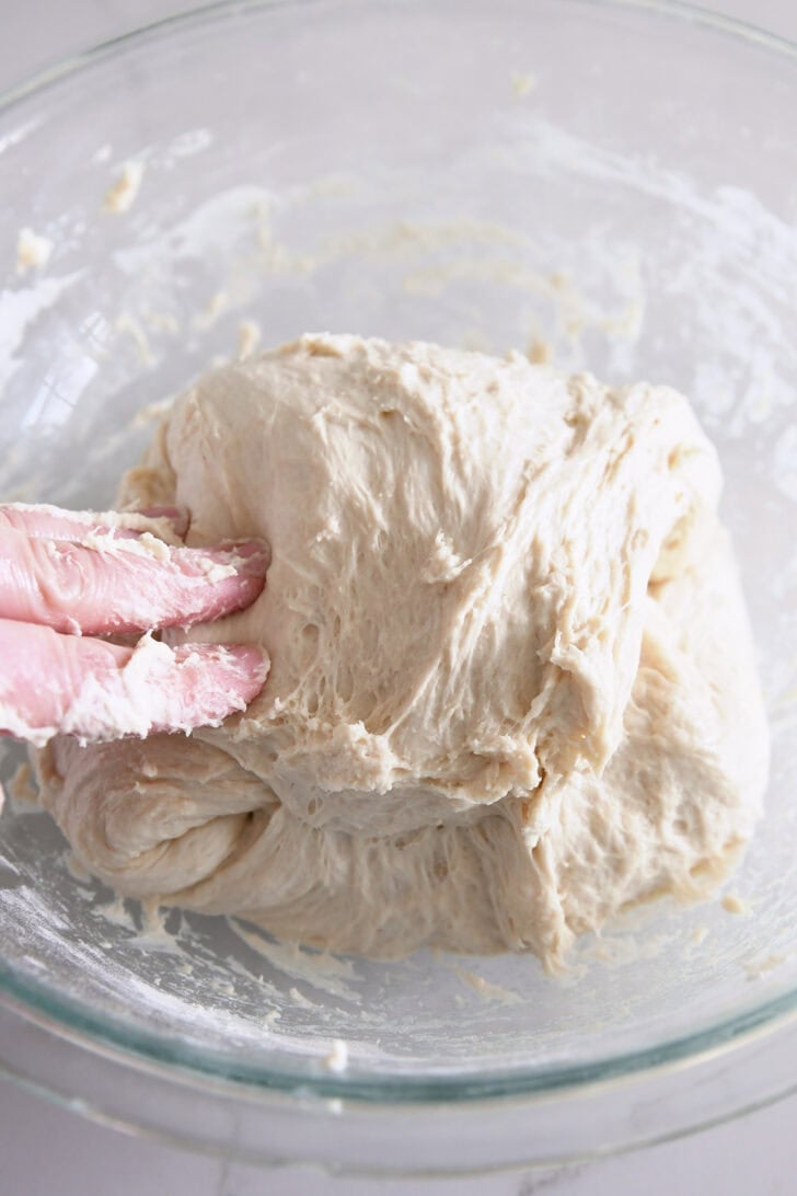 Folding ciabatta dough over itself in glass bowl.