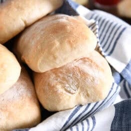 Several baked and flour dusted ciabatta rolls in white dish with blue and white towel.