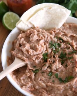 White bowl with creamy refried beans, cilantro, tortillas and wooden spoon.