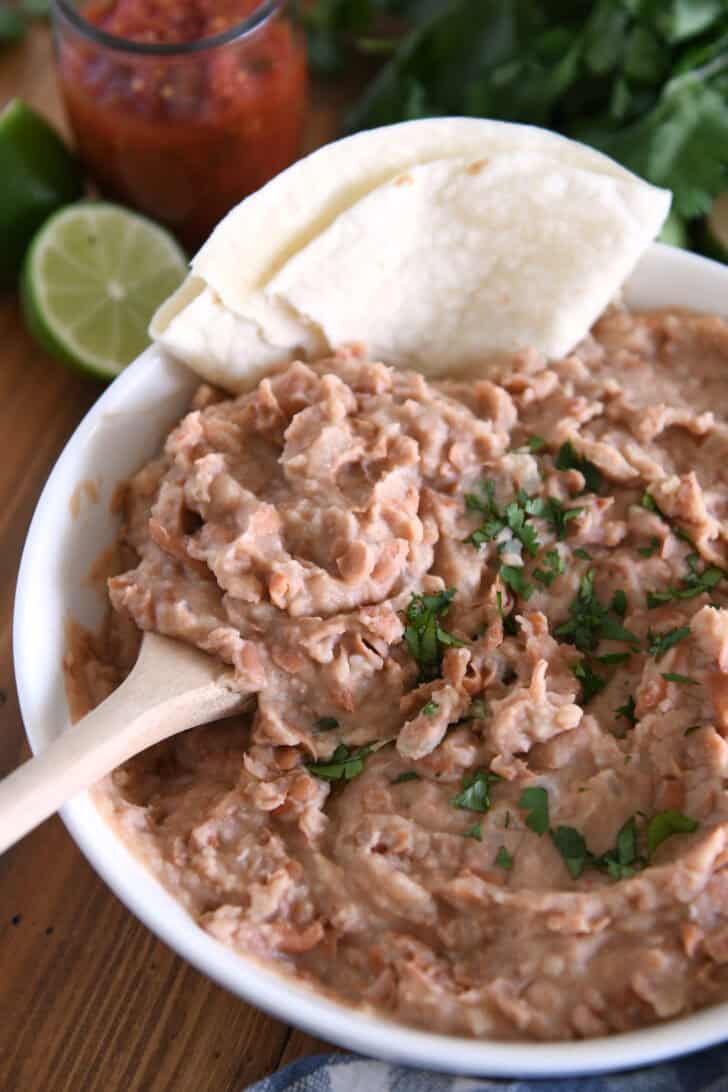 White bowl with creamy refried beans, cilantro, tortillas and wooden spoon.