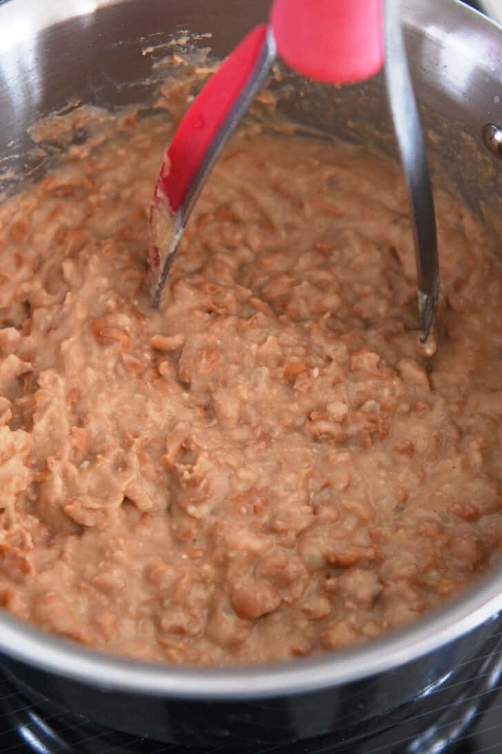 Potato masher mashing canned beans in stainless pot.