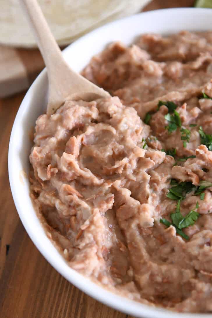 Wooden spoon scooping serving of refried beans from white bowl.