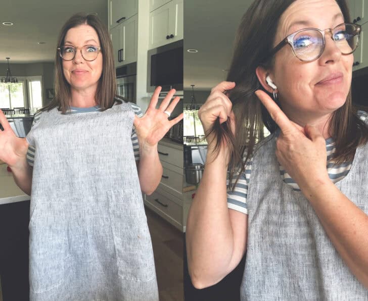Woman in apron in kitchen pointing to earbuds in ears. 