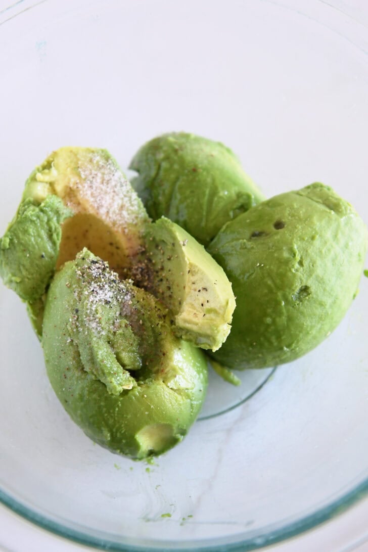 Four halved avocados with salt, lime juice and pepper in glass bowl.