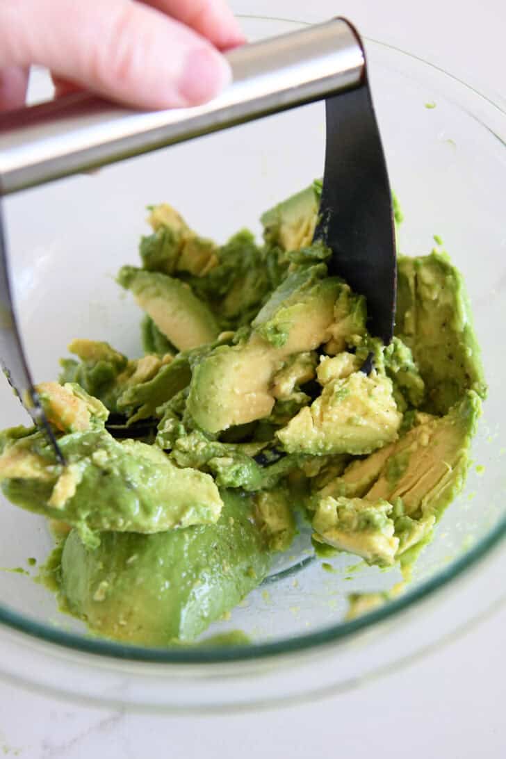 Using pastry blender to mash avocados in glass bowl.