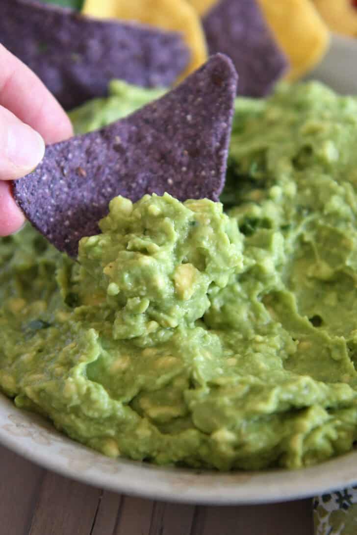 Blue tortilla chip scooping up homemade guacamole in tan bowl.