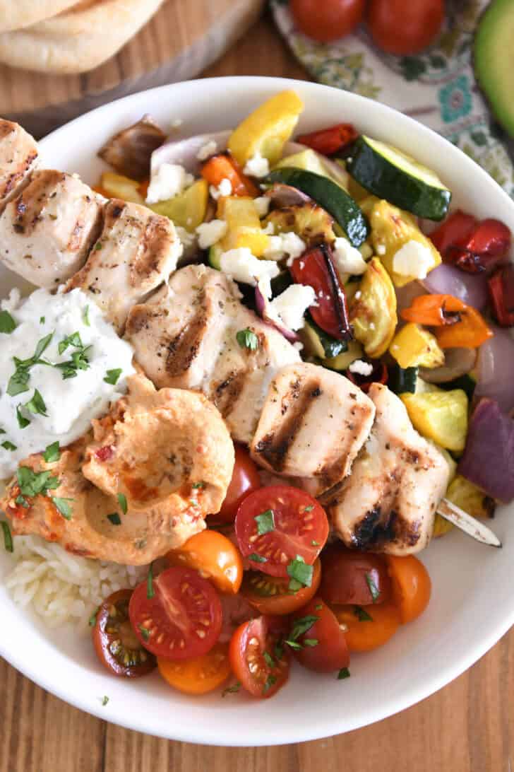 Top down view of white bowl with metal skewer with chicken pieces, roasted squash, zucchini, red onions and bell peppers, cherry tomatoes, rice, hummus and tzatziki sauce.