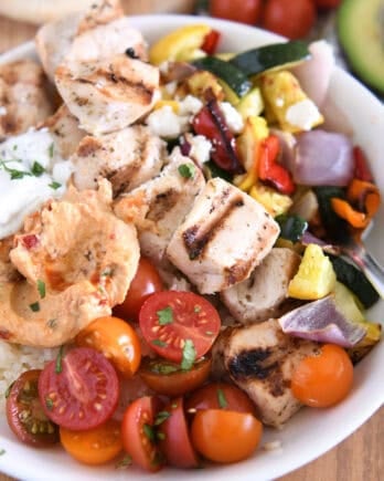 White bowl with metal skewer with chicken pieces, roasted squash, zucchini, red onions and bell peppers, cherry tomatoes, rice, hummus and tzatziki sauce.