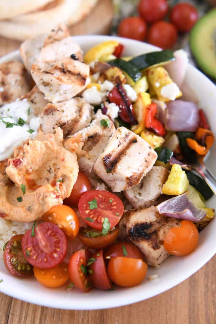 White bowl with metal skewer with chicken pieces, roasted squash, zucchini, red onions and bell peppers, cherry tomatoes, rice, hummus and tzatziki sauce.