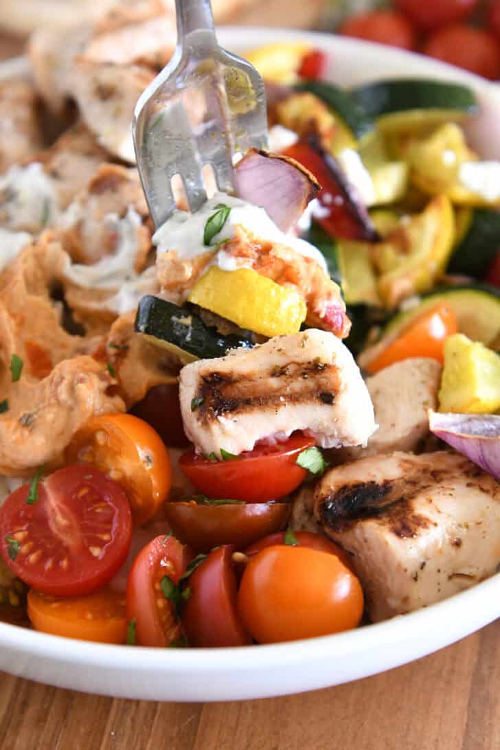 Fork piercing bite of chicken, roasted veggies hummus and tzatziki.