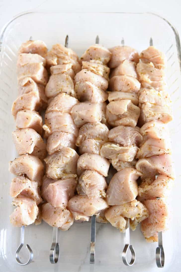 Glass 9X13-inch dish with skewers of raw chicken in lemon marinade.