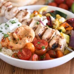 White bowl with metal skewer with chicken pieces, roasted squash, zucchini, red onions and bell peppers, cherry tomatoes, rice, hummus and tzatziki sauce.