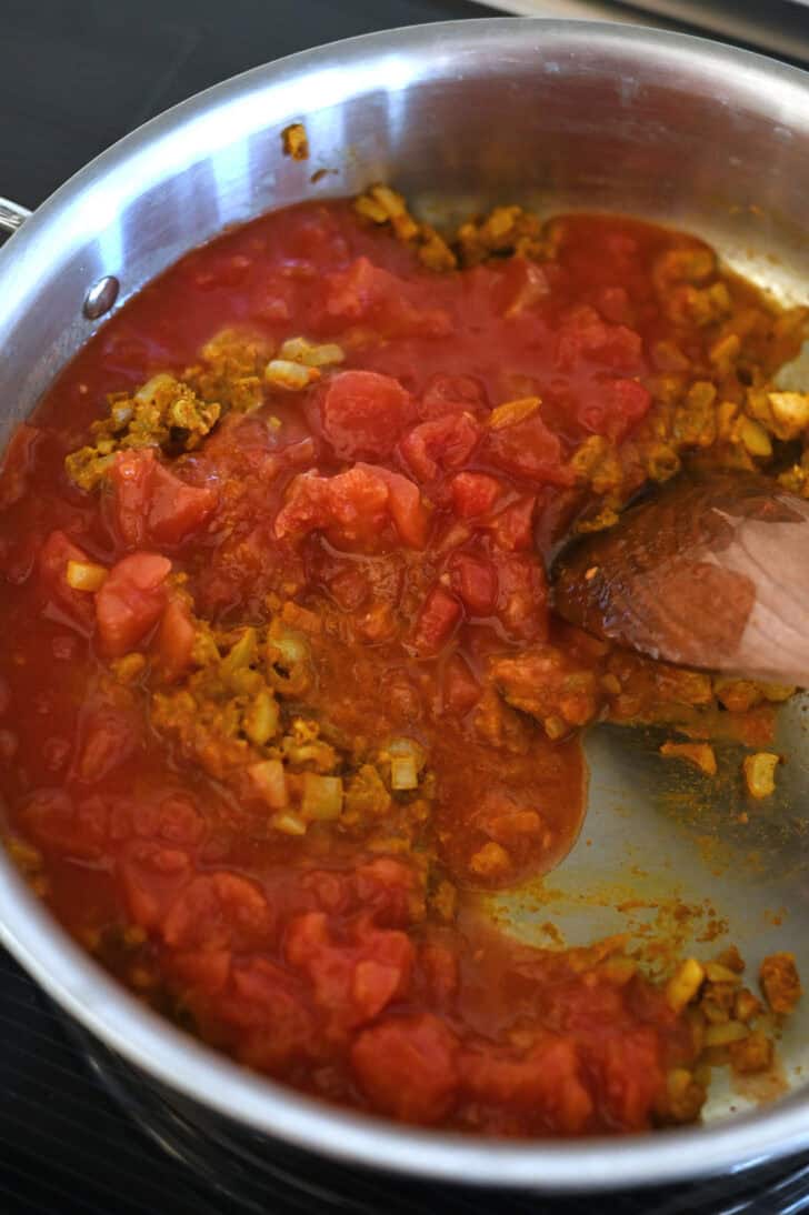 Diced tomatoes, curry spices, and onions cooking in a metal pot.