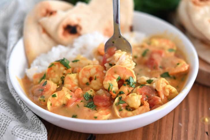 A fork taking a bite of rice and shrimp curry out of a white bowl with flatbread on the side.