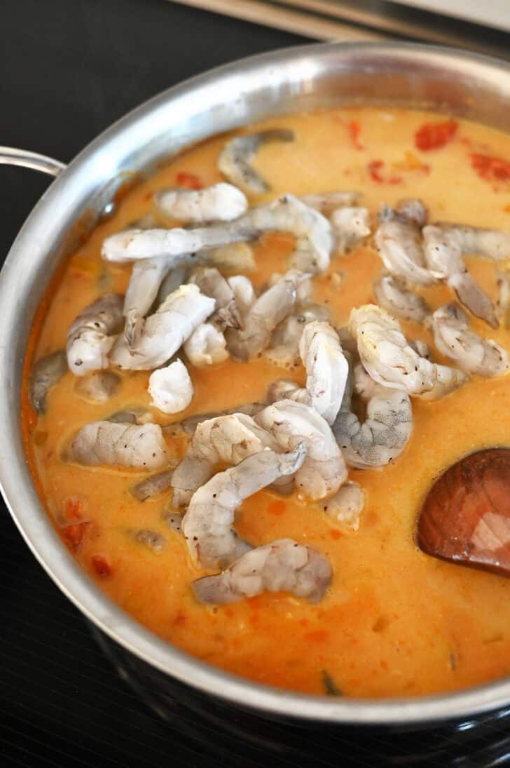 Raw shrimp going into a metal pot with a tomato curry sauce.