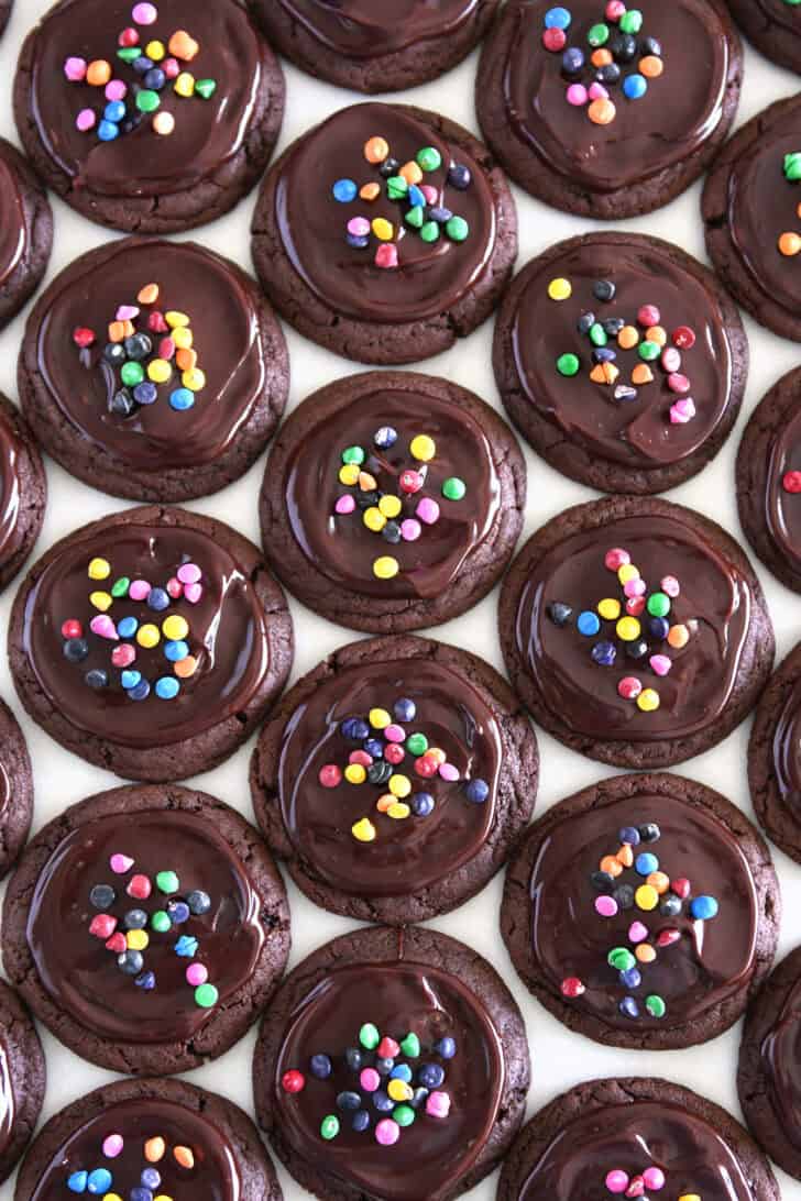 Top down view of many chocolate cookies topped with ganache and sprinkled with rainbow chips.