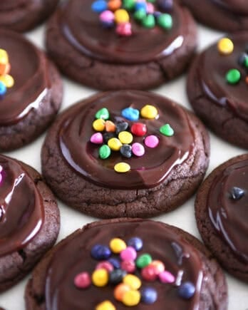 Several frosted chocolate cookies on white parchment paper topped with rainbow chips.