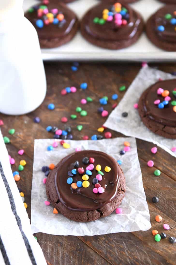 Chocolate cookie topped with ganache and rainbow chips on white parchment paper with rainbow chips sprinkled around and several cookies in the background.