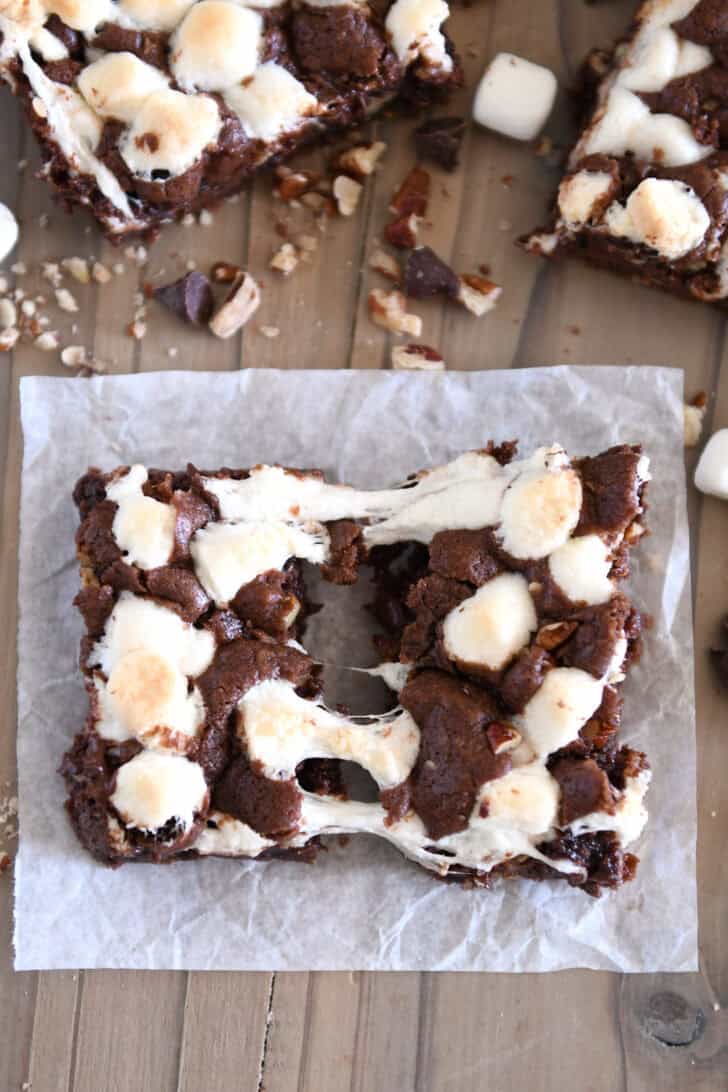 One chocolate cookie bar with pecans and marshmallows pulled apart with stretchy marshmallows on square spatula on white parchment paper.