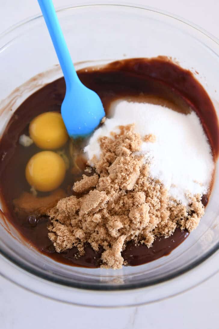 Glass bowl with melted chocolate, eggs, granulated sugar, brown sugar and blue silicone spatula.
