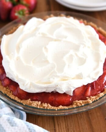 A strawberry pie with graham cracker crust in glass pie plate with whipped cream on top.