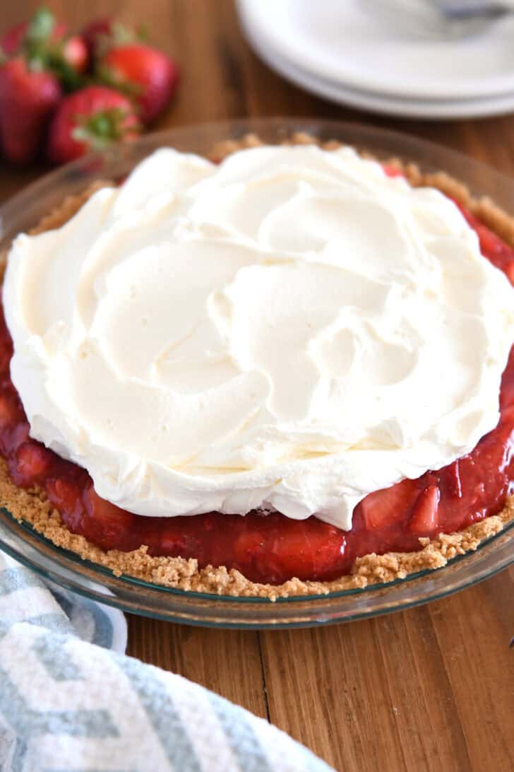 A strawberry pie with graham cracker crust in glass pie plate with whipped cream on top.