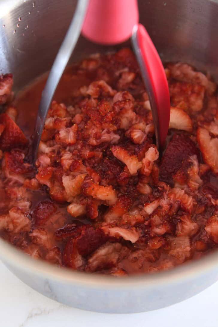 Potato masher mashing fresh strawberries in stainless pot.