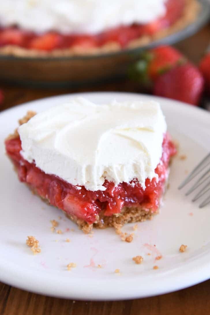 Half eaten slice of strawberry pie with whipped cream on top on white plate.