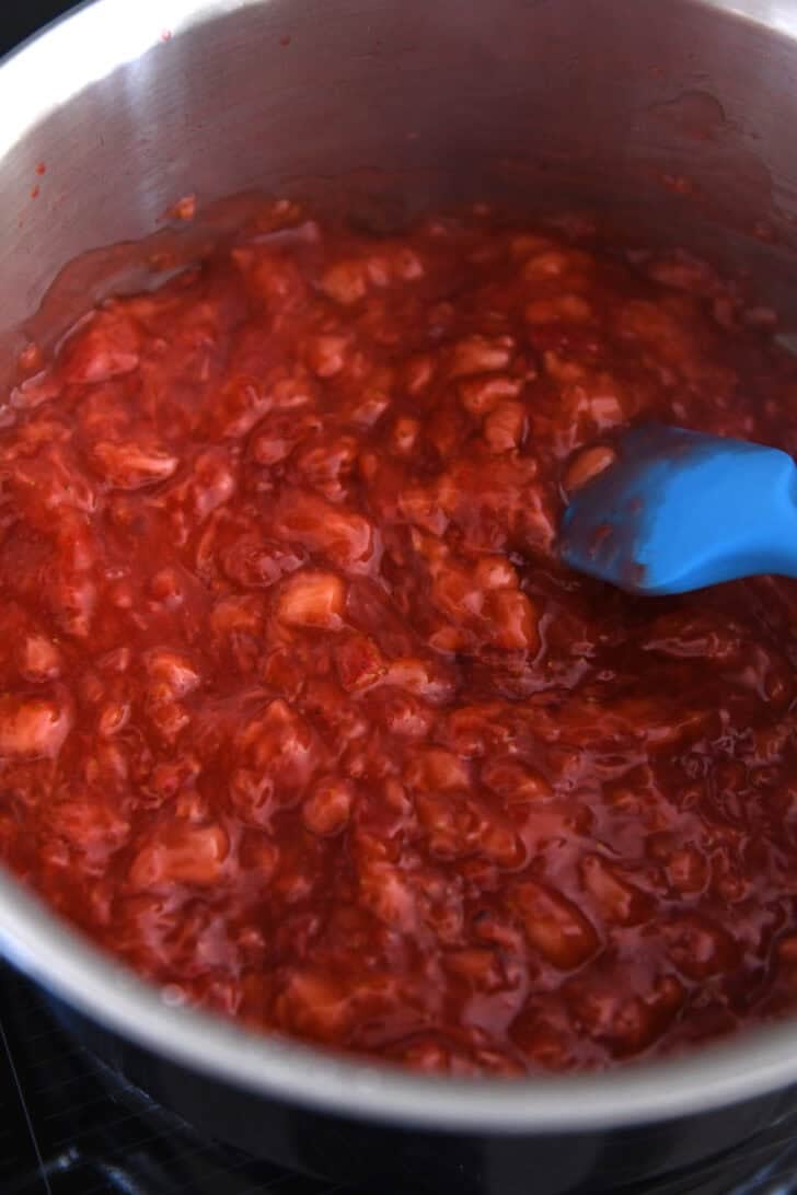 Strawberry mixture simmering on stove in stainless pot with blue spatula.