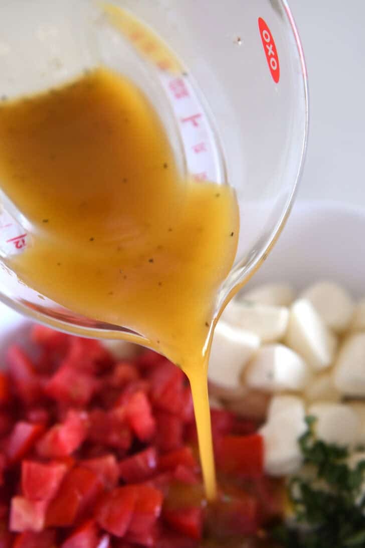 A glass measuring cup pouring an oil mixture into a white dish with chopped tomatoes and mozzarella.
