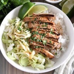 Top down photo of white bowl with chopped cabbage, white rice, grilled chicken, lime wedges, chopped cilantro and toasted sesame seeds.