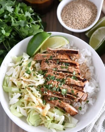 Top down photo of white bowl with chopped cabbage, white rice, grilled chicken, lime wedges, chopped cilantro and toasted sesame seeds.