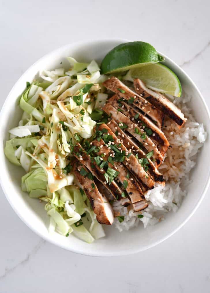 White bowl with chopped cabbage, white rice, grilled chicken, cilantro and lime wedges.