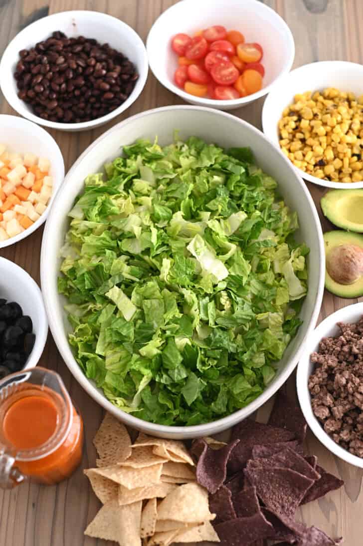 Bowl of lettuce in the center with bowls of black beans, chopped tomatoes, roasted corn, cubed cheese, avocados, and cooked ground beef on the side.