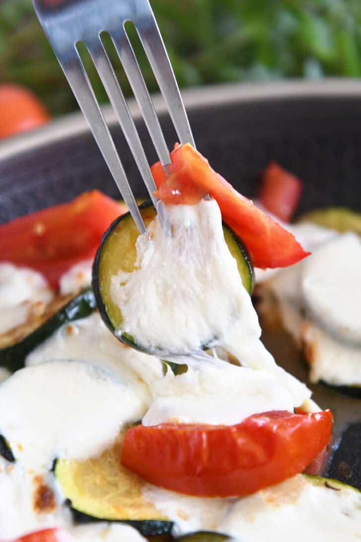 A fork is holding a piece of sautéed zucchini with melted mozzarella and tomato.