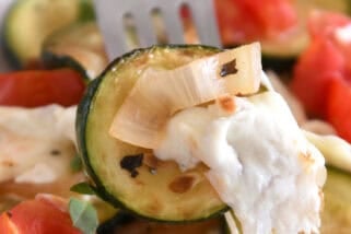 A fork is holding a piece of sautéed zucchini with melted mozzarella, tomatoes, and sautéed onions.