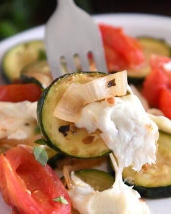 A fork is holding a piece of sautéed zucchini with melted mozzarella, tomatoes, and sautéed onions.