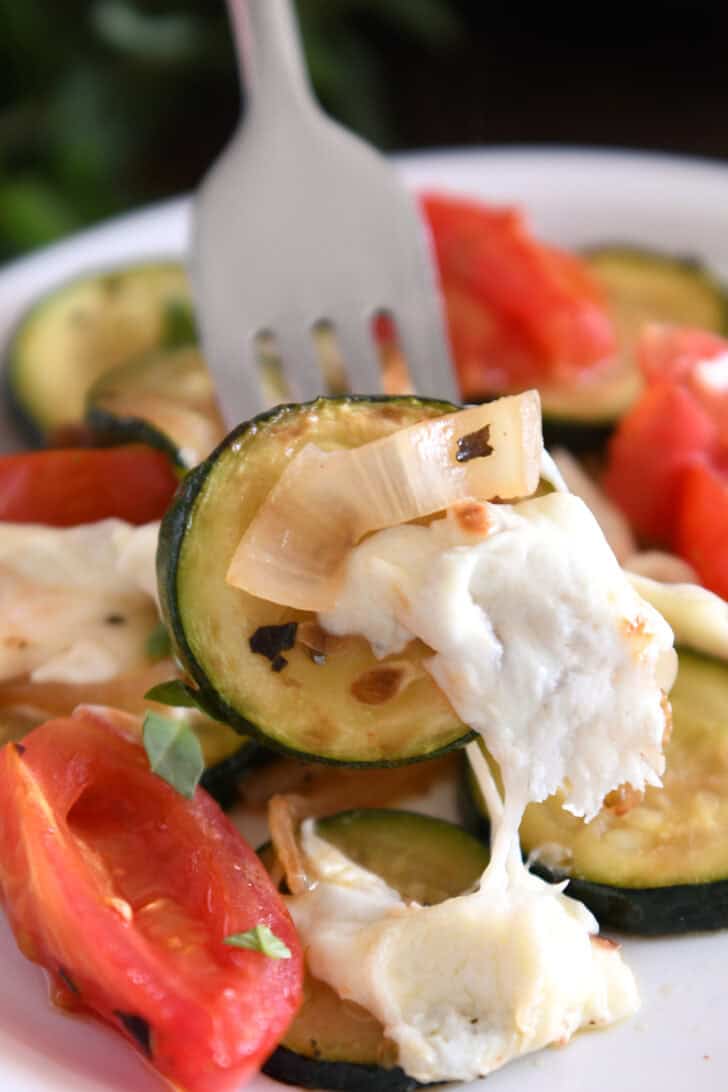 A fork is holding a piece of sautéed zucchini with melted mozzarella, tomatoes, and sautéed onions.