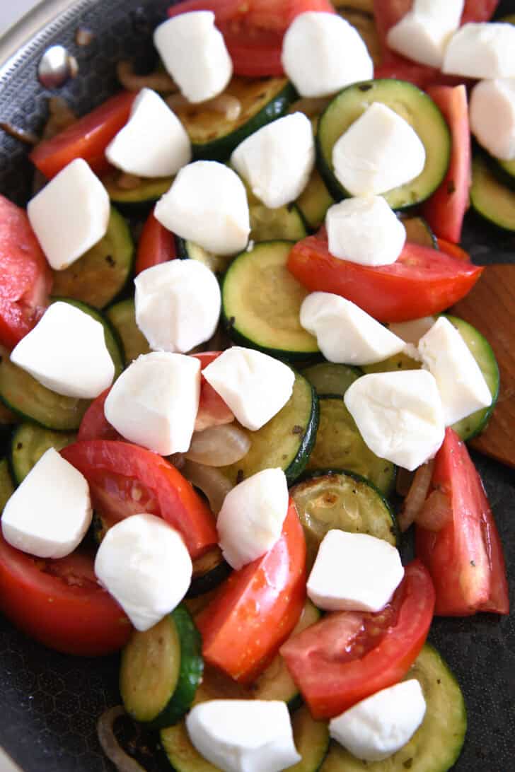 Top down view of a skillet full of sautéed zucchini and tomatoes with chunks of fresh mozzarella on top of the vegetables.