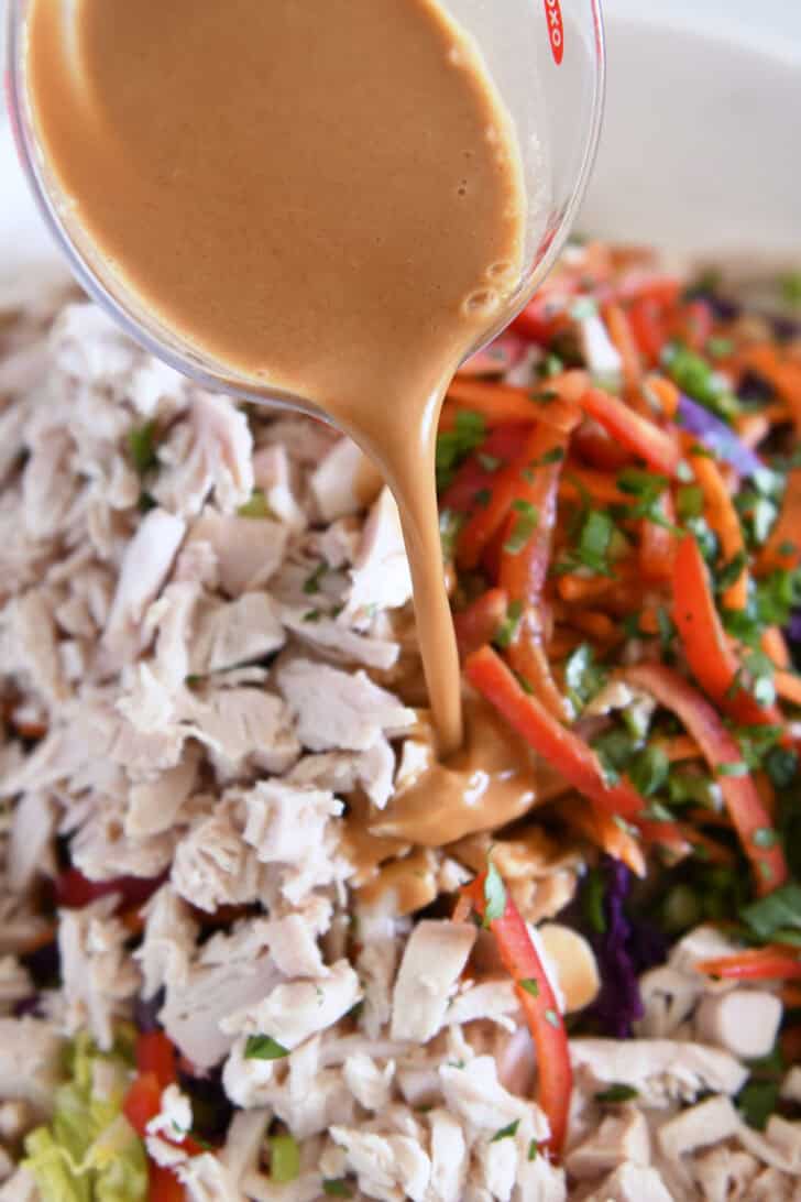 Pouring peanut vinaigrette over ingredients for Asian chicken salad in white bowl.