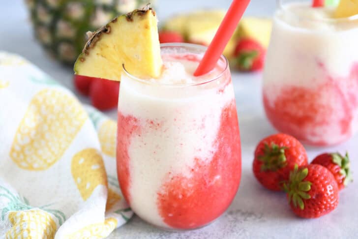 Glass cup filled with strawberry puree and icy pineapple coconut mixture with red straw and pineapple wedge on rim of glass.