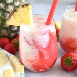 Glass cup filled with strawberry puree and icy pineapple coconut mixture with red straw and pineapple wedge on rim of glass.
