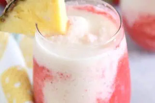 Glass cup filled with strawberry puree and icy pineapple coconut mixture with red straw and pineapple wedge on rim of glass.