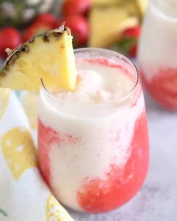 Glass cup filled with strawberry puree and icy pineapple coconut mixture with red straw and pineapple wedge on rim of glass.