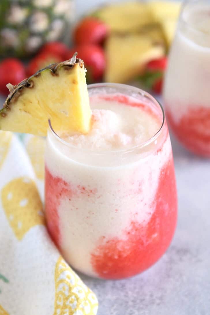 Glass cup filled with strawberry puree and icy pineapple coconut mixture with red straw and pineapple wedge on rim of glass.