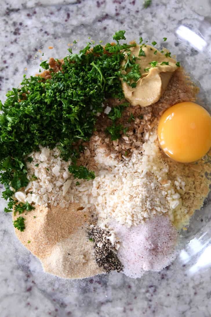 Top down view of a raw egg, spices, and chopped parsley in a glass bowl.